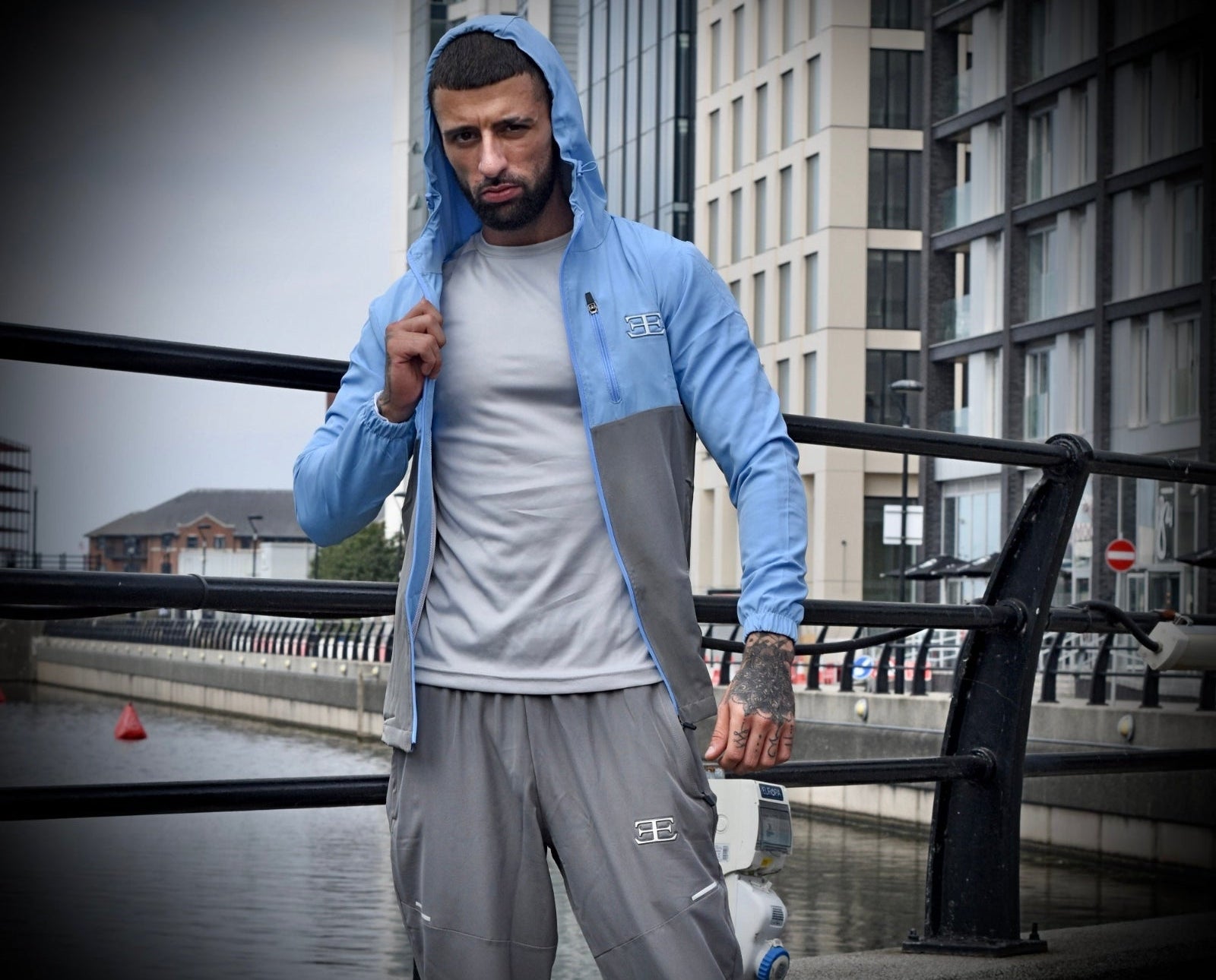 man wearing light blue tracksuit with hood up with Elegante clothing logo on chest and grey tracksuit joggers, building in the background