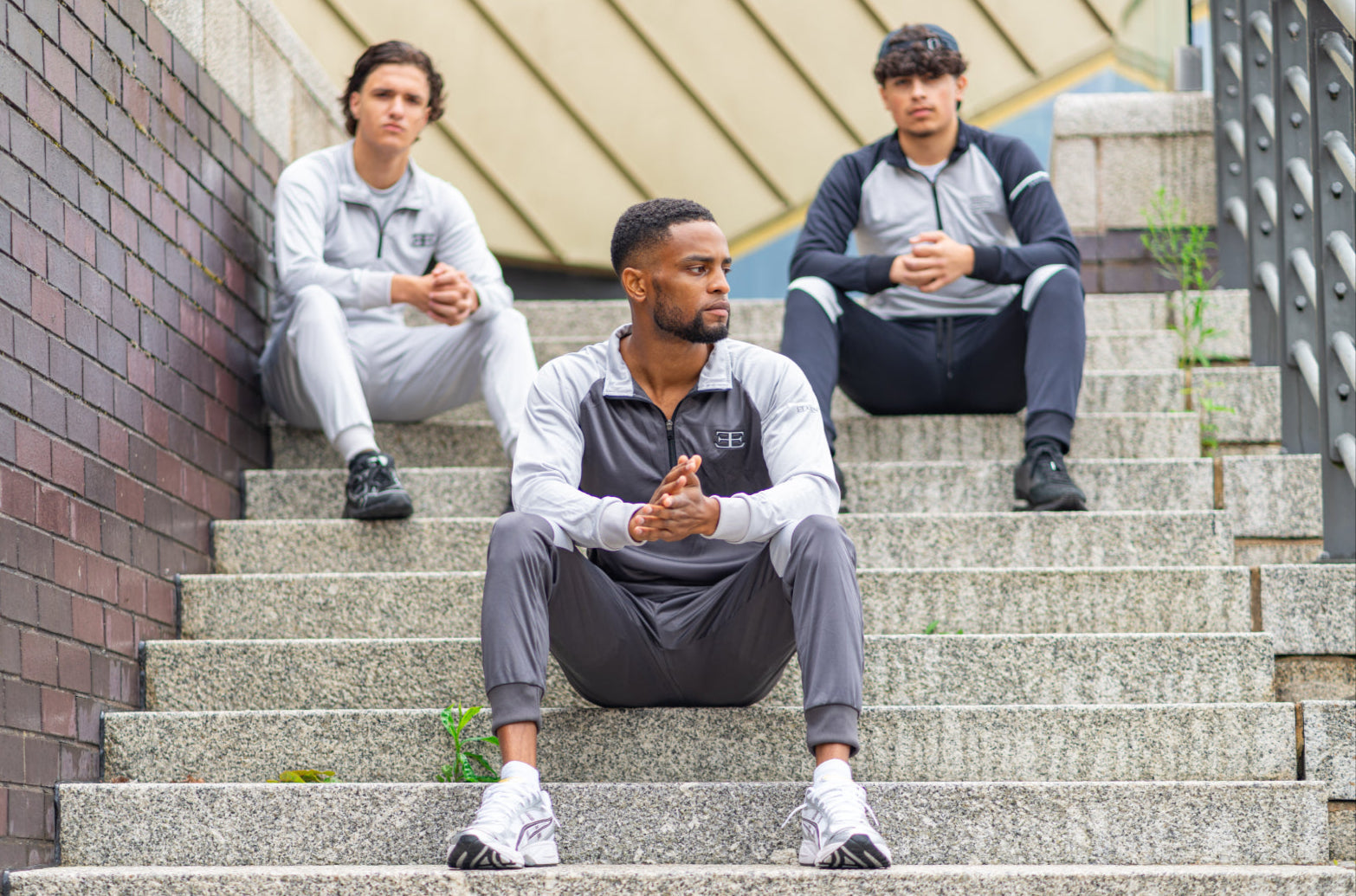 3 Man wearing Grey Tracksuits with Elegante Logo on chest sat on steps