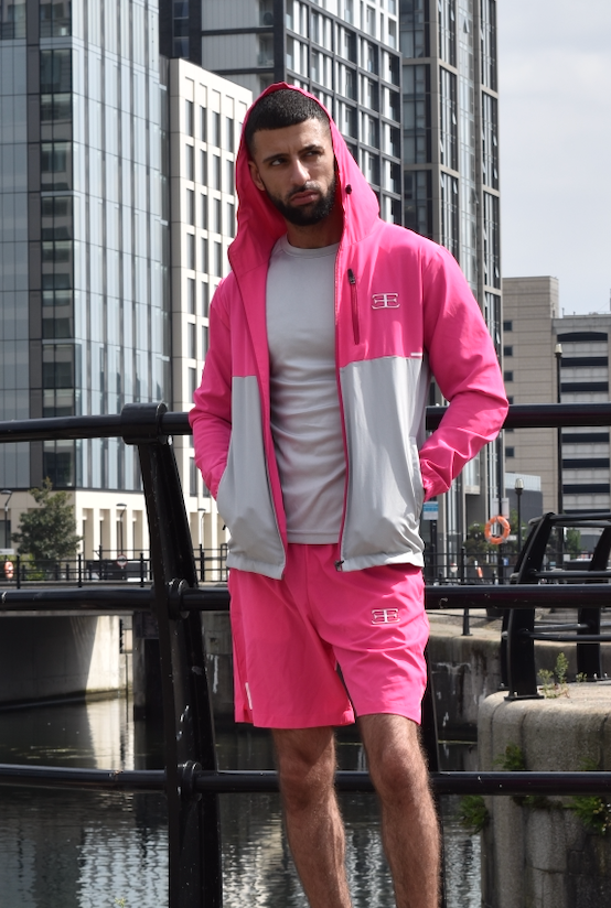 man wearing pink tracksuit jacket with hood up and pink shorts with buildings in background