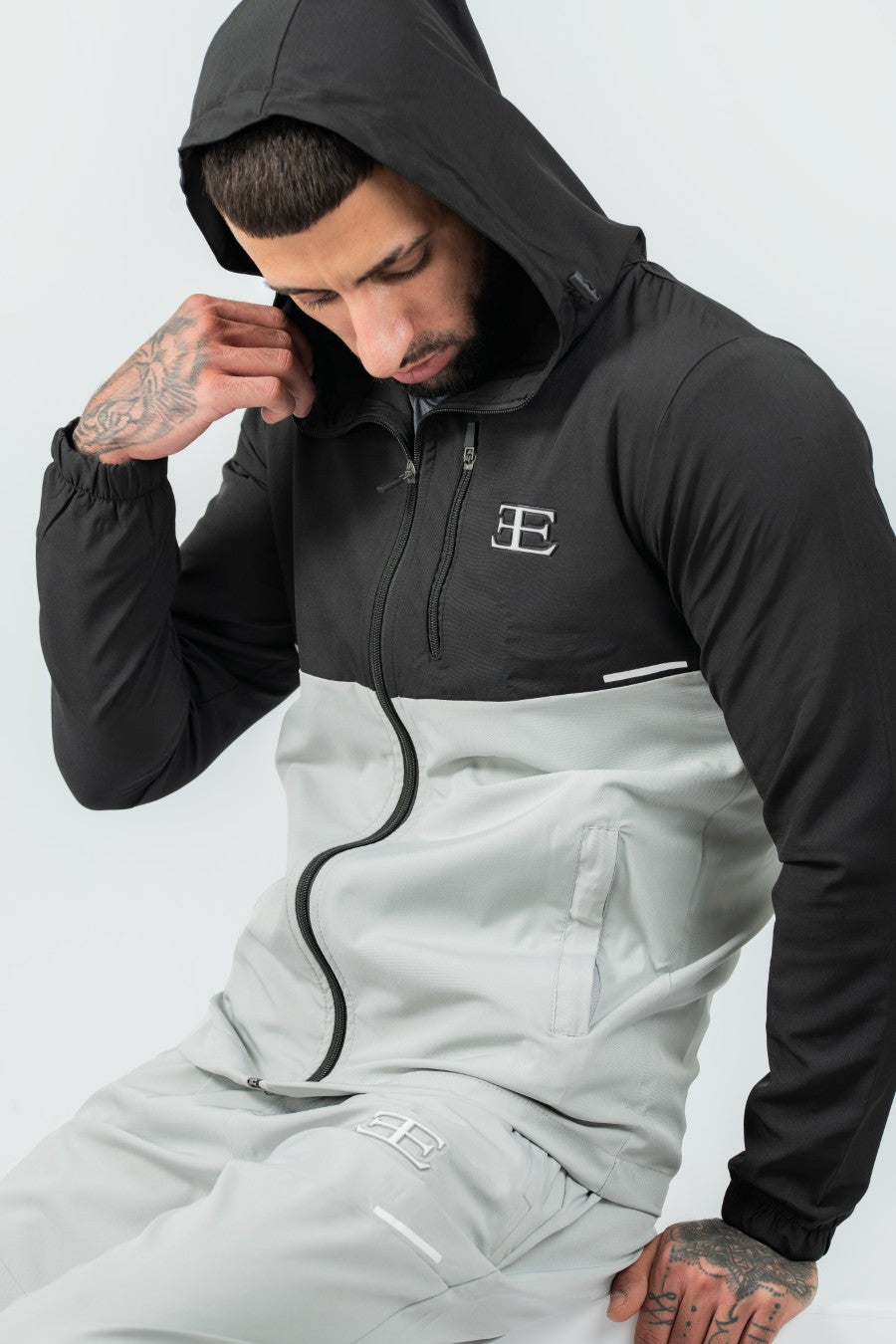 man wearing a gray and black tracksuit jacket and joggers with hood up on a white background
