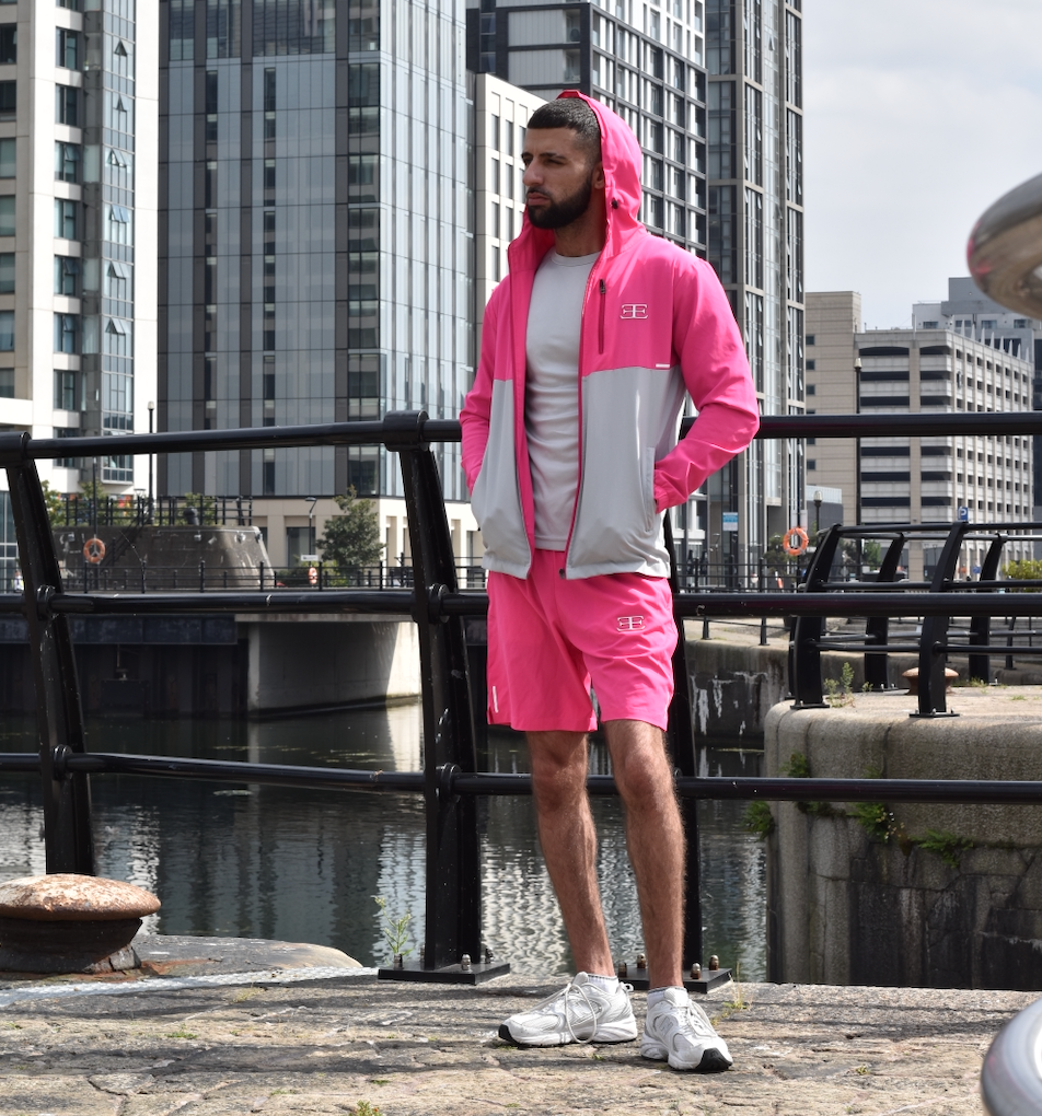 Man wearing a pink tracksuit standing by a waterfront with buildings in the background